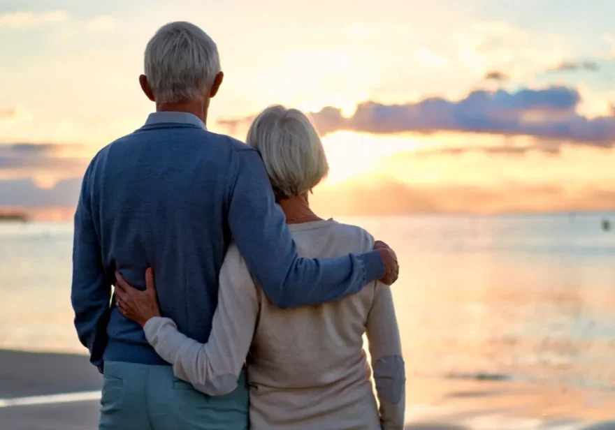 An elderly couple embracing while watching a sunset over the ocean.